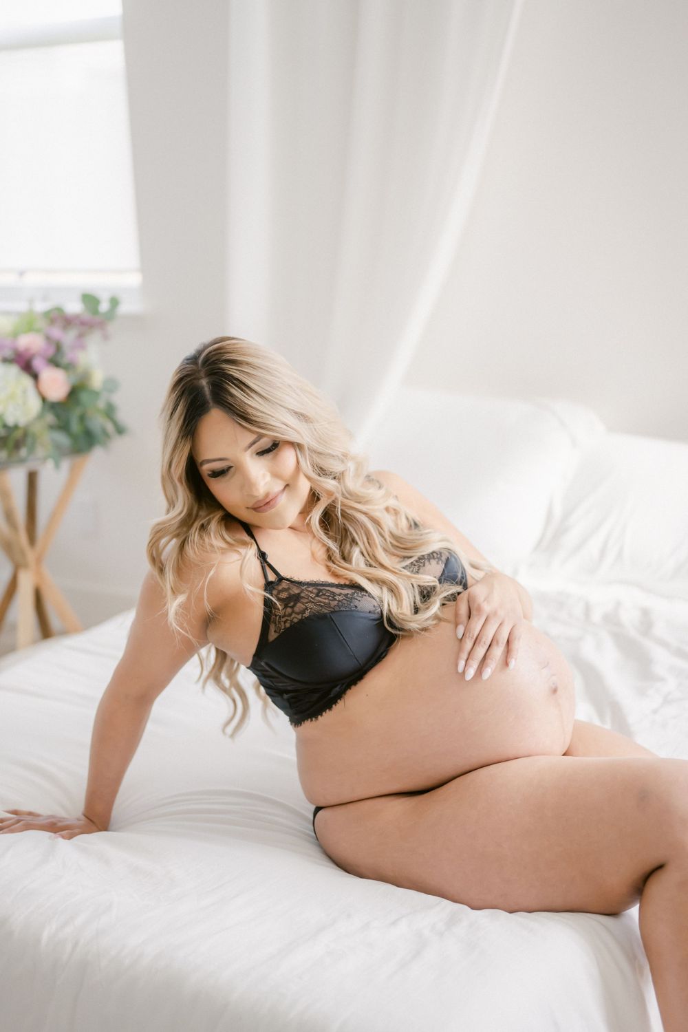 Expectant woman in black lingerie poses on white bedding in soft natural window light during maternity photoshoot.