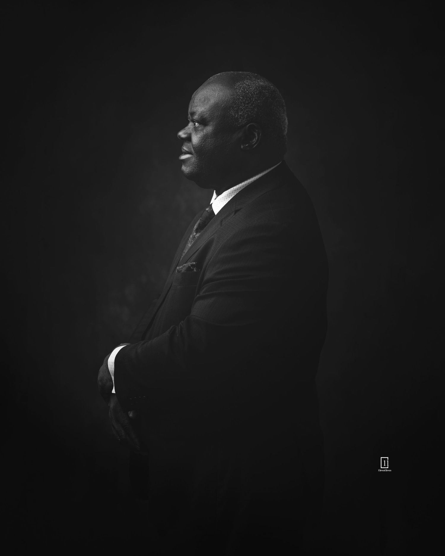 Dramatic black and white portrait showing a profile view of a figure in a suit against a dark background.