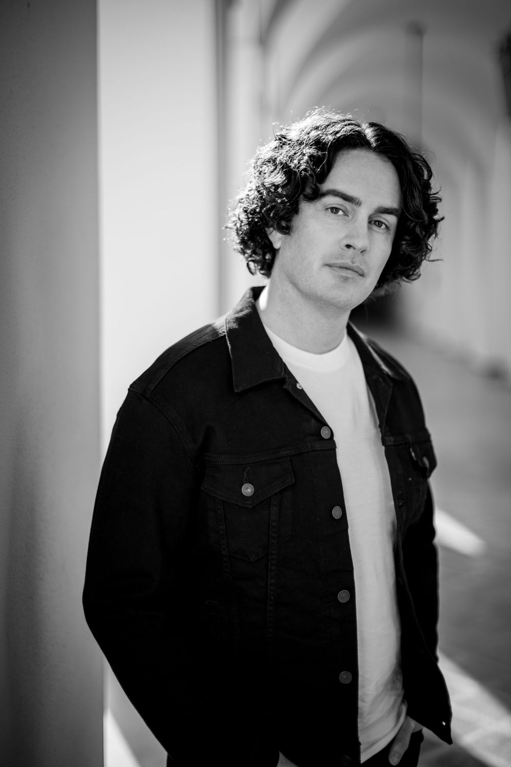 Black and white portrait of a person with dark curly hair wearing a denim jacket and white shirt against a light background.