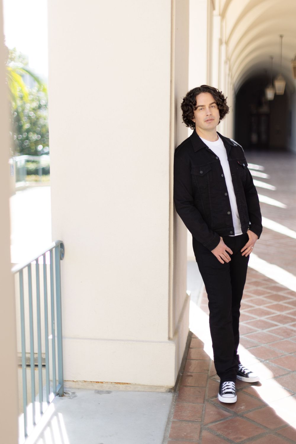 A figure stands casually against a white column in an arched hallway wearing black jeans and a jacket over a white shirt.
