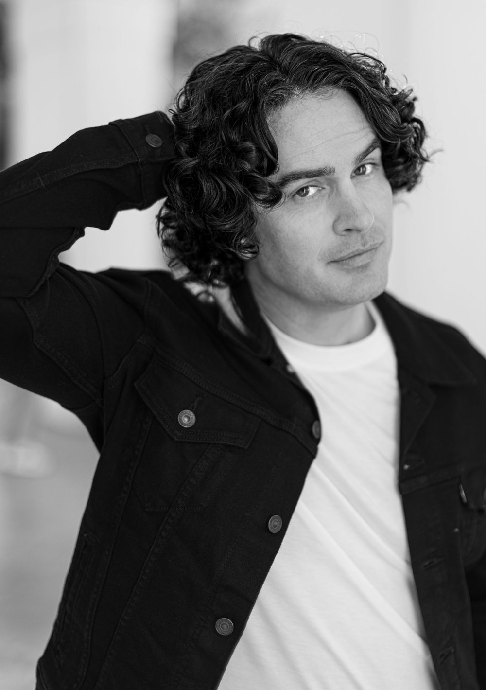 Black and white portrait of a person with dark curly hair wearing a white t-shirt and dark jacket.