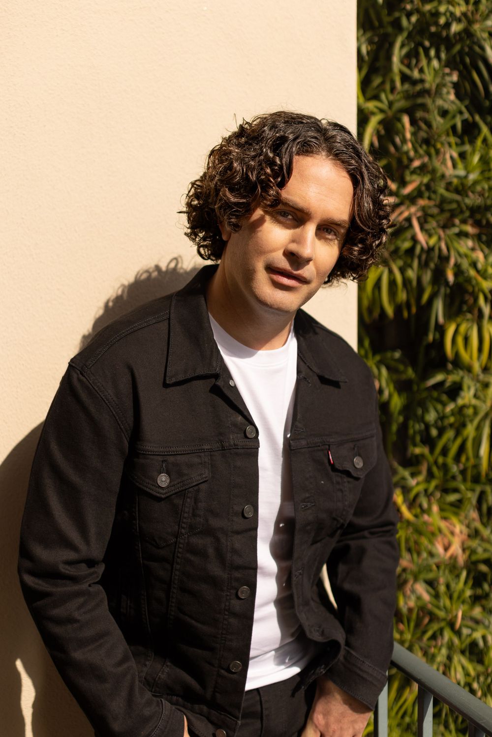 Person in dark denim jacket and white t-shirt poses against beige wall with greenery in background.