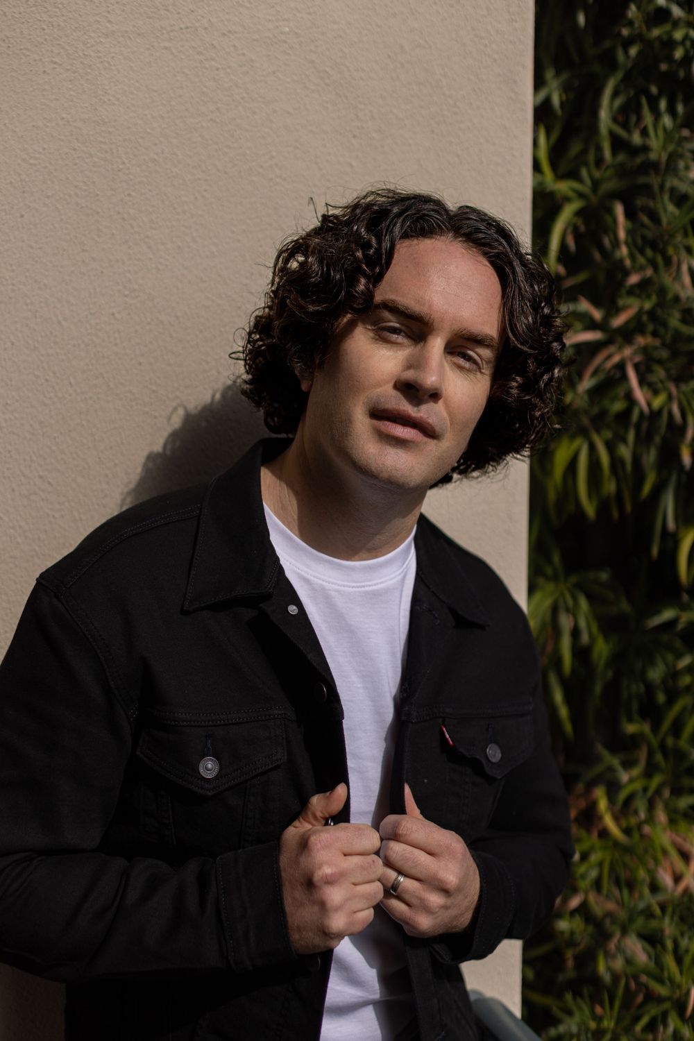 Portrait against beige wall wearing white t-shirt and black jacket in natural sunlight.