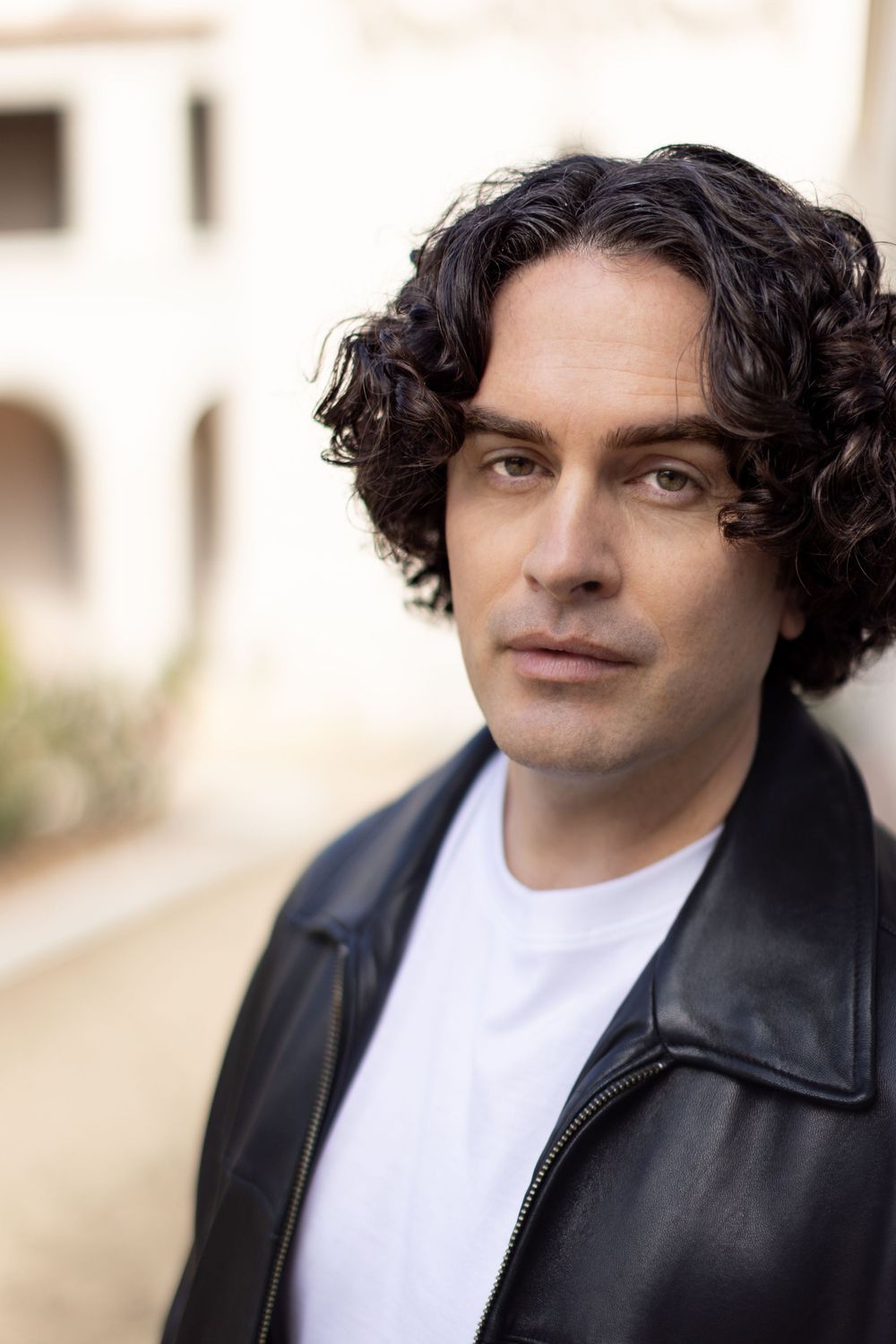 A person with dark curly hair wearing a black leather jacket and white t-shirt poses outdoors against a blurred background.