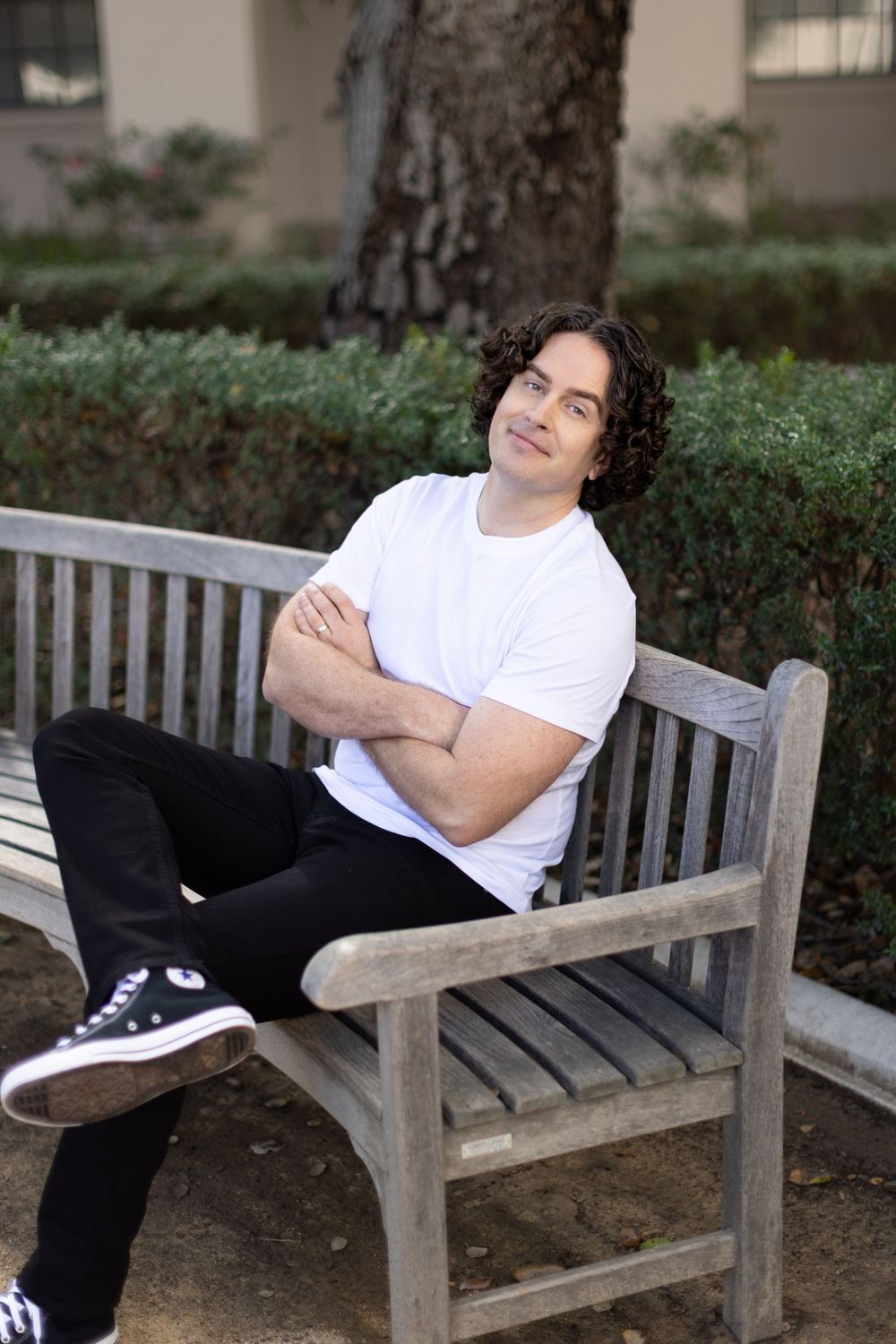 Person relaxes on a wooden bench outdoors wearing a white t-shirt and black pants against a garden background.