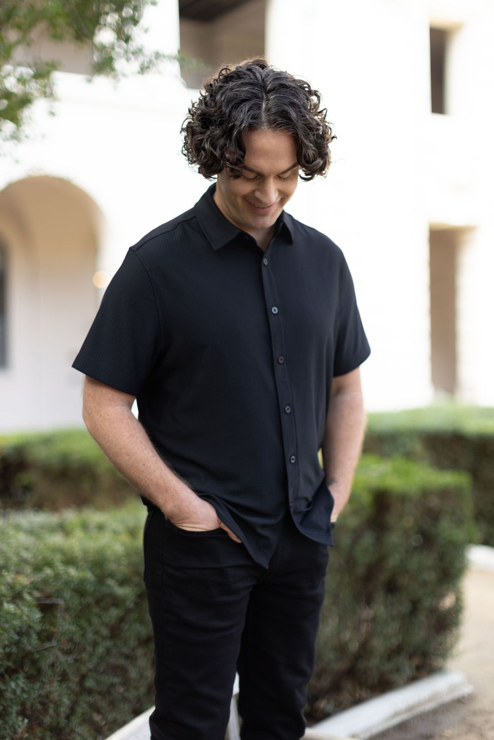 Casual portrait of someone wearing a black short-sleeve button down shirt with curly dark hair looking down.