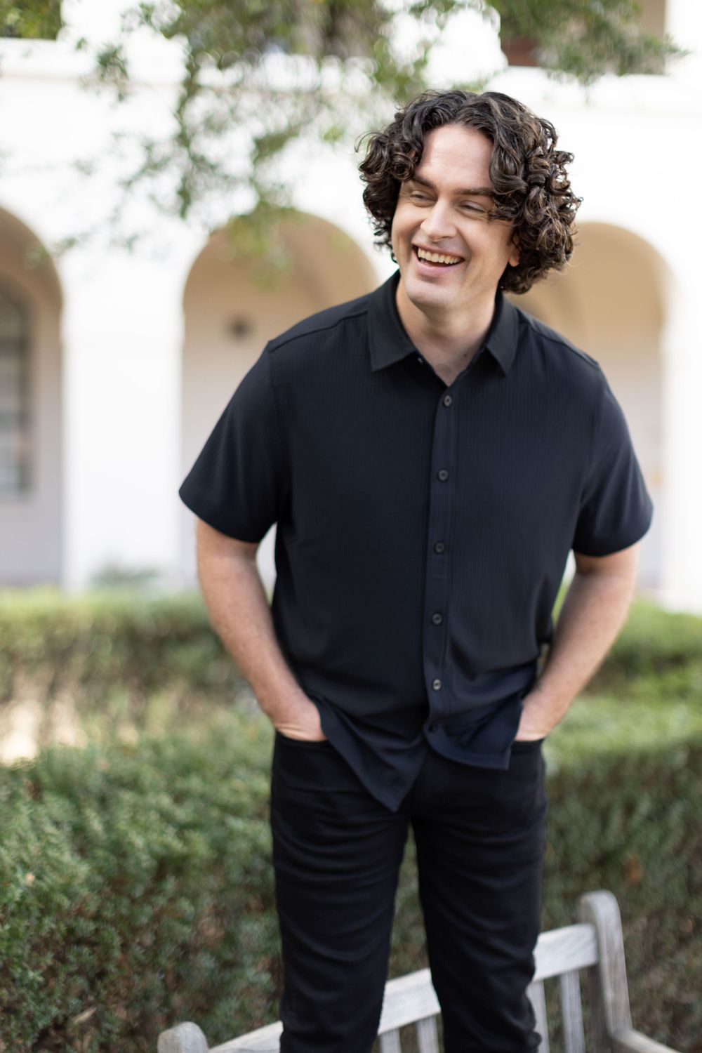 Person in a black casual button-down shirt and dark pants stands smiling against an architectural backdrop.
