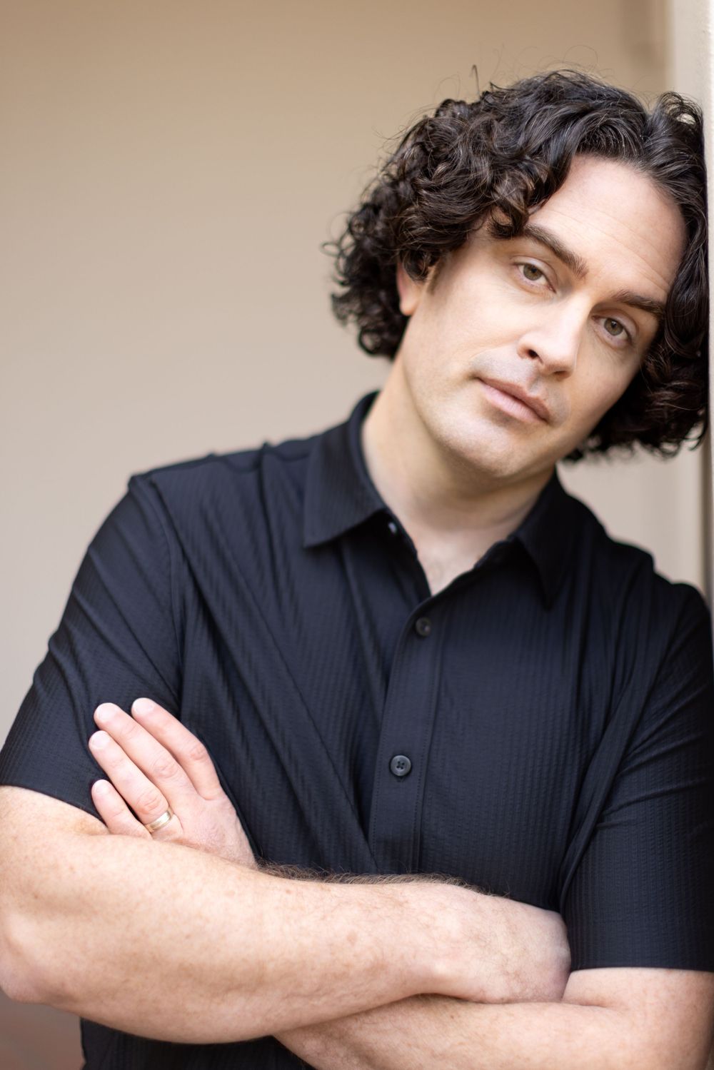 Portrait of person with dark curly hair wearing a black polo shirt against a light wall.