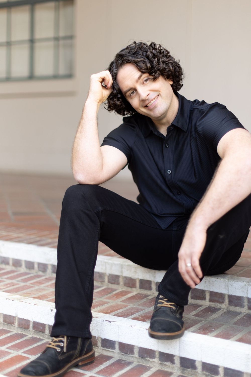 Casual outdoor portrait sitting on brick steps wearing all black attire with curly hair.