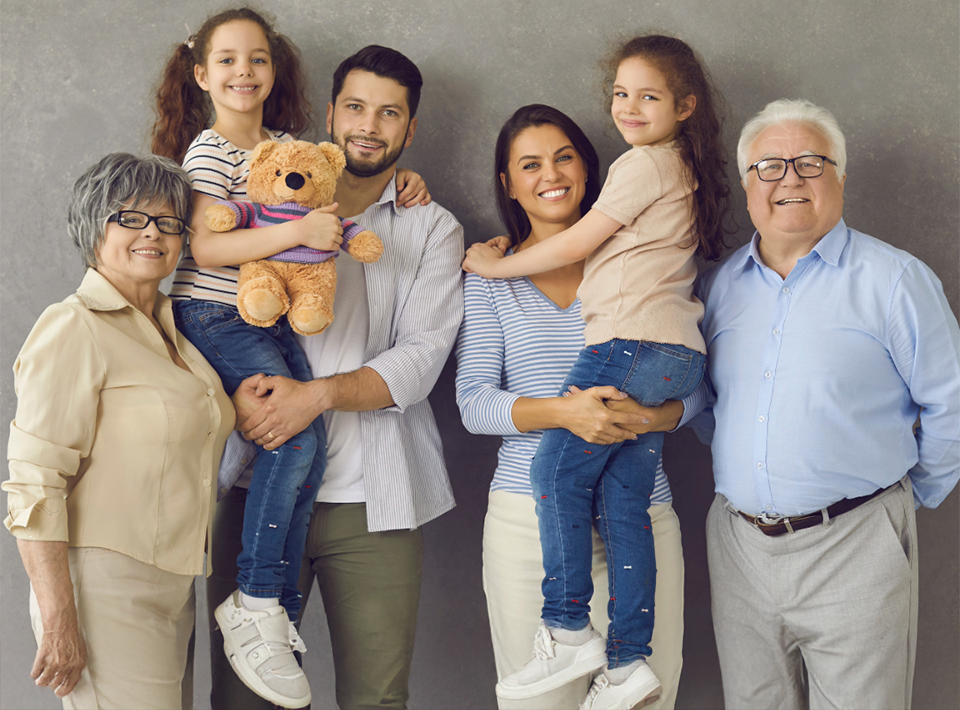 Multi-generational group posing together with teddy bear on gray background.