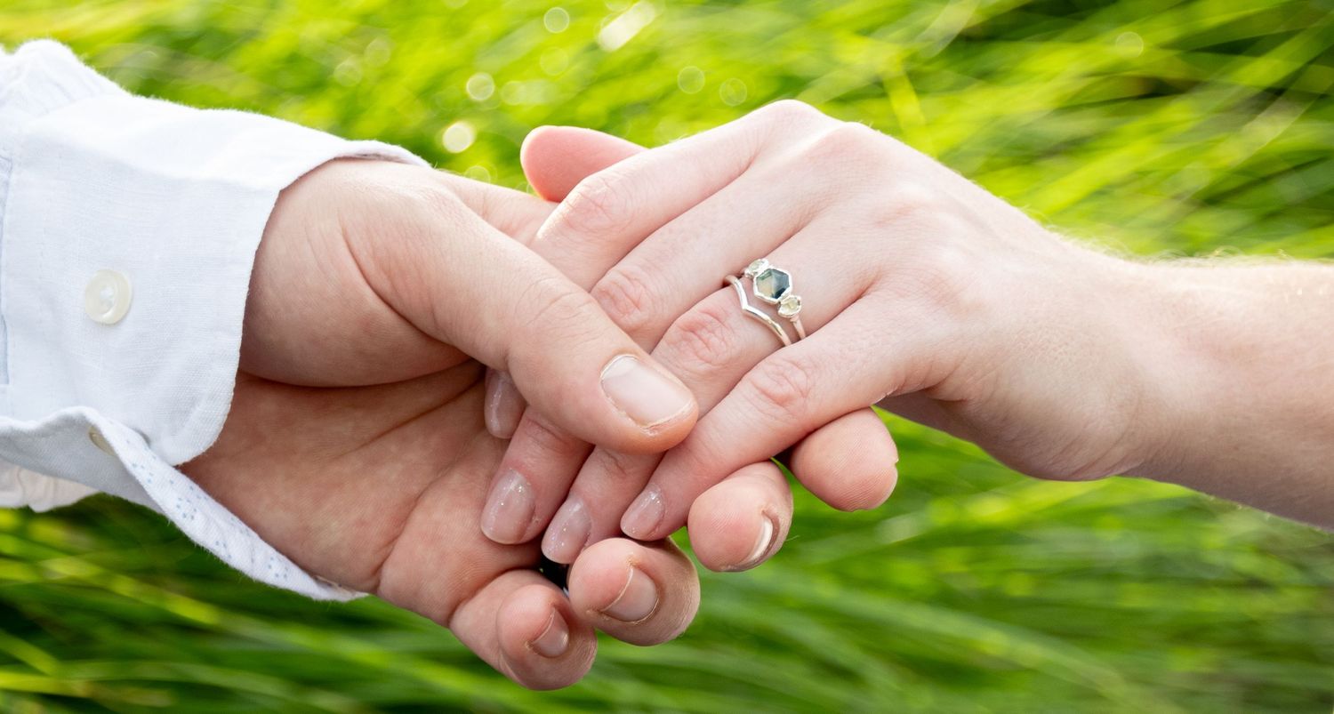Close-up photo of two hands holding each other with an engagement ring outdoors against blurred green background.