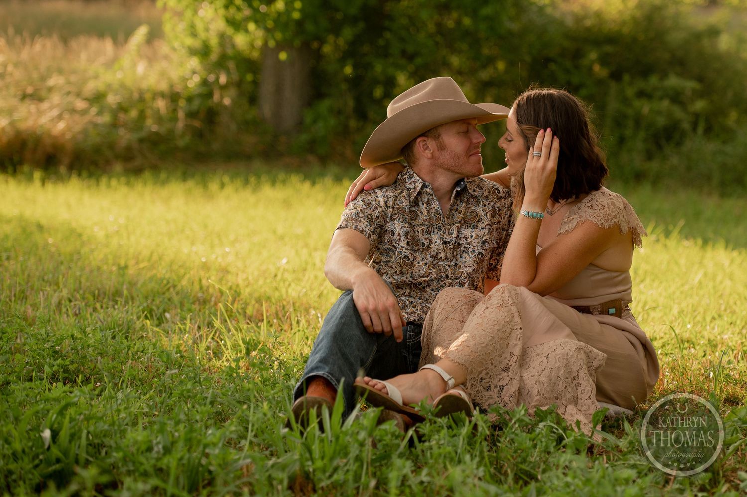 A tender moment between a couple sitting in a grassy field at sunset wearing western attire.