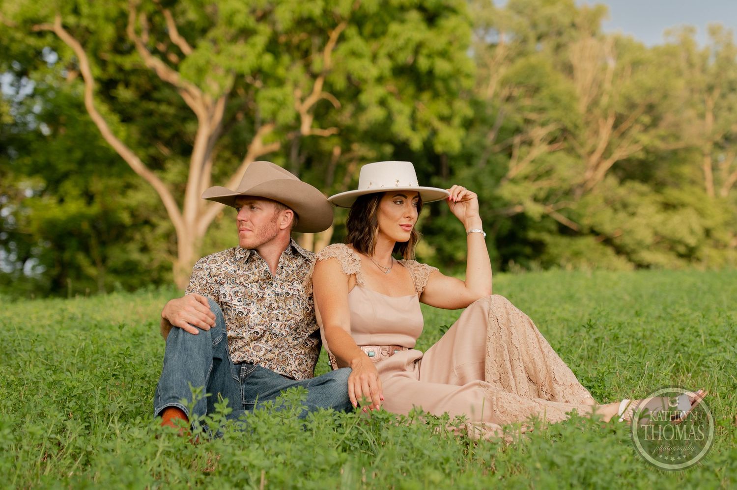 A couple in western wear relaxing on a grassy hill with trees in the background.