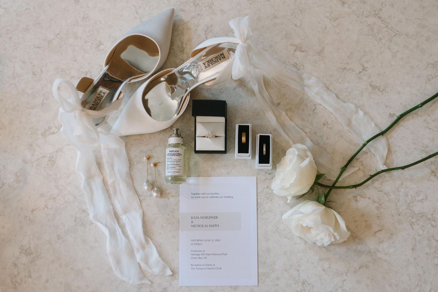 Wedding details including rings, jewelry boxes, delicate ribbon, white roses, and invitation paper laid out on light marble surface.
