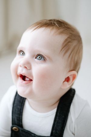 A happy baby with bright eyes and a joyful smile wearing a white shirt and dark overalls.