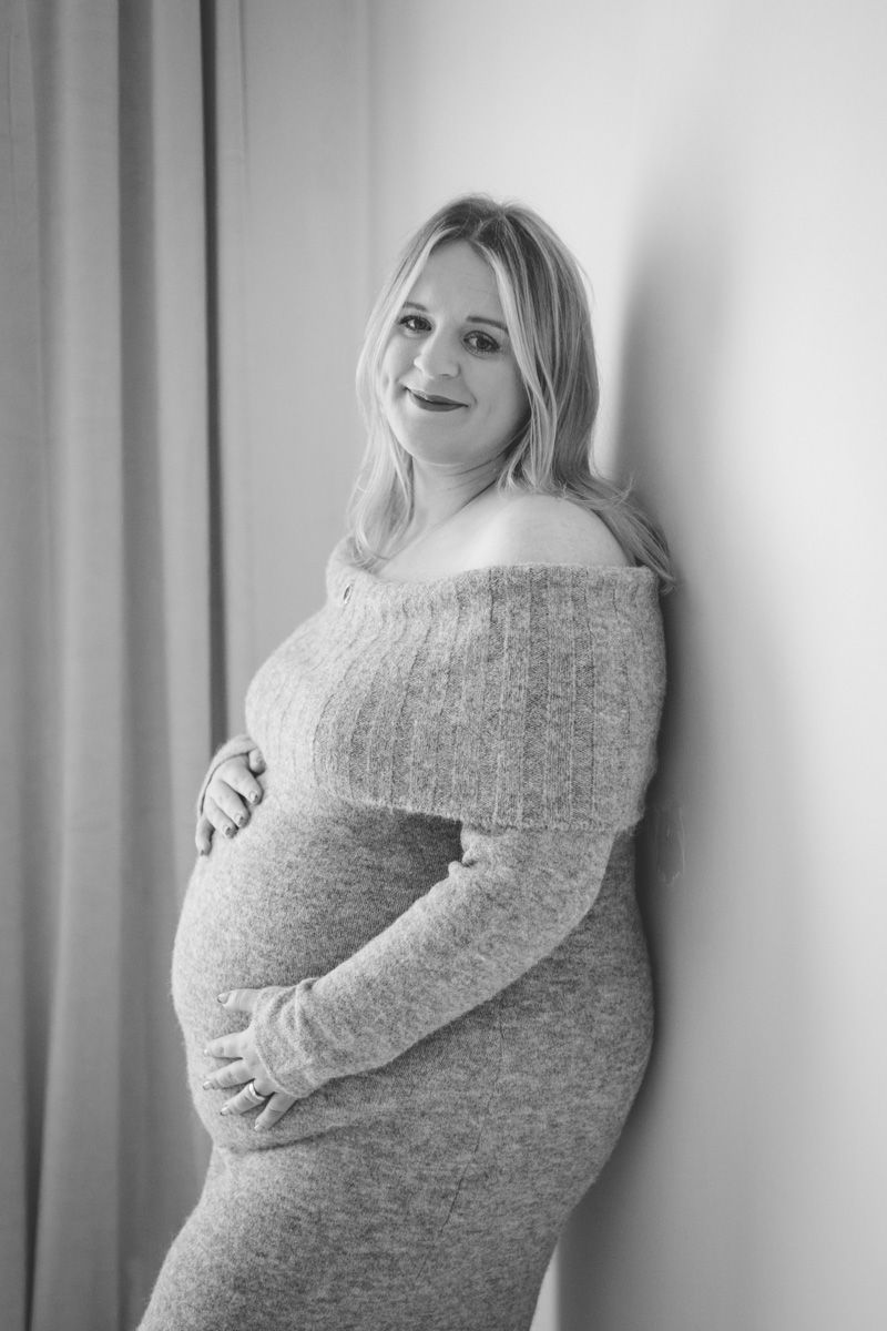Black and white maternity photo series showing pregnant woman in cozy sweater cradling baby bump against striped wall.