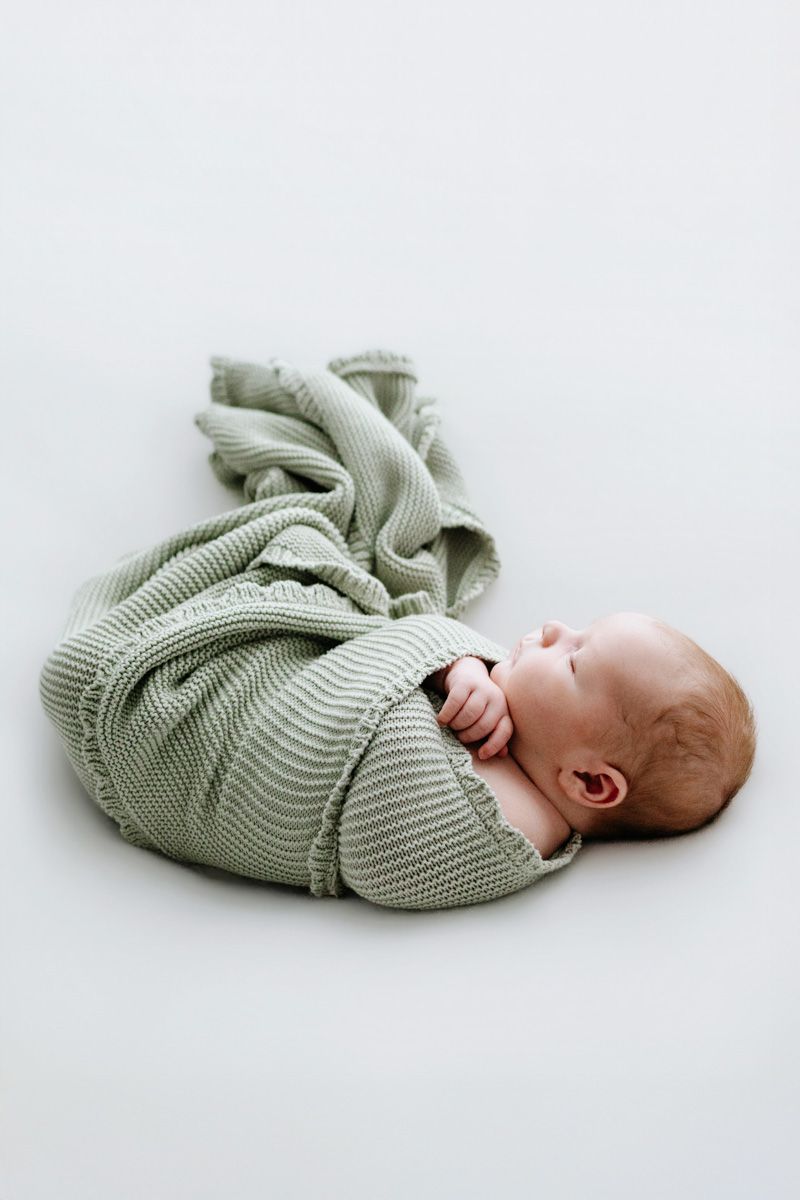 Newborn baby swaddled in a soft sage green knit blanket, lying peacefully on a white background.