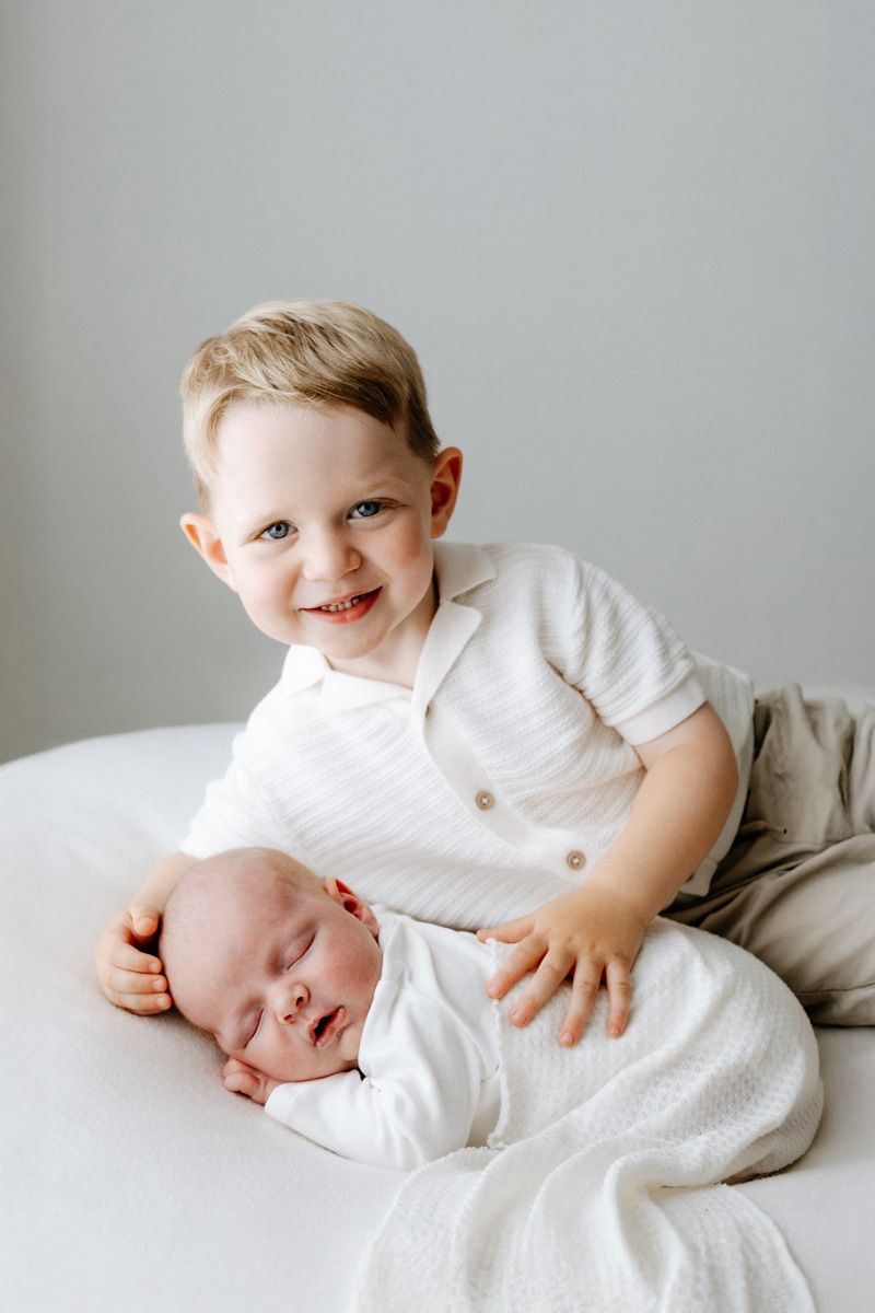 Smiling toddler boy in white shirt poses with sleeping newborn baby sibling on white surface.