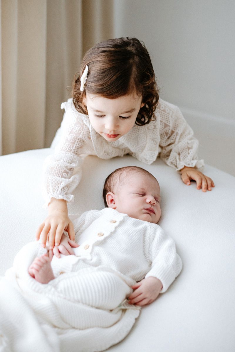 A tender moment captured in white as a child in lace gently embraces a sleeping newborn on soft bedding.