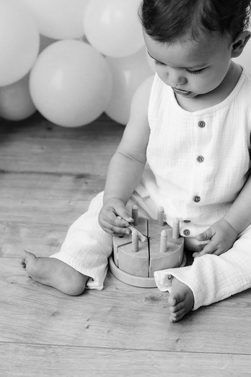 Black and white photo of a baby sitting on a wooden floor playing with a wooden toy, with balloons in the background.