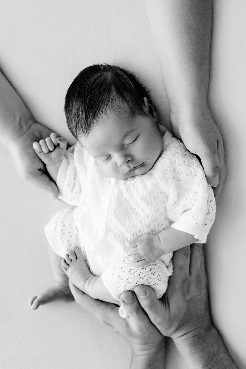 Black and white newborn photo of sleeping baby in white lace outfit cradled gently in parents hands.