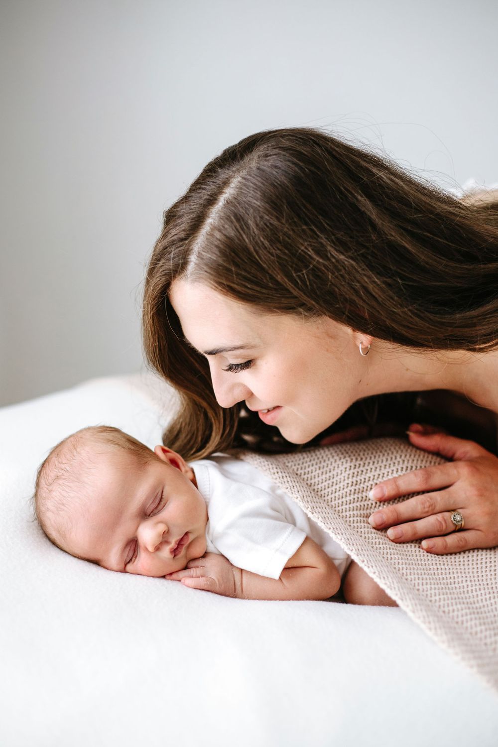 Mother sweetly gazes at her sleeping newborn baby while lying together on white bedding.