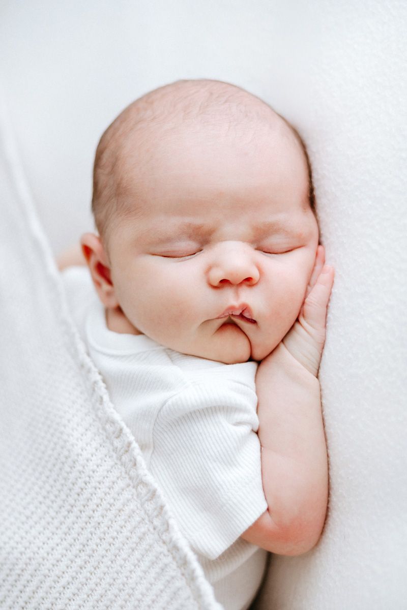 A sleeping newborn baby in white clothing rests peacefully on white textured bedding.