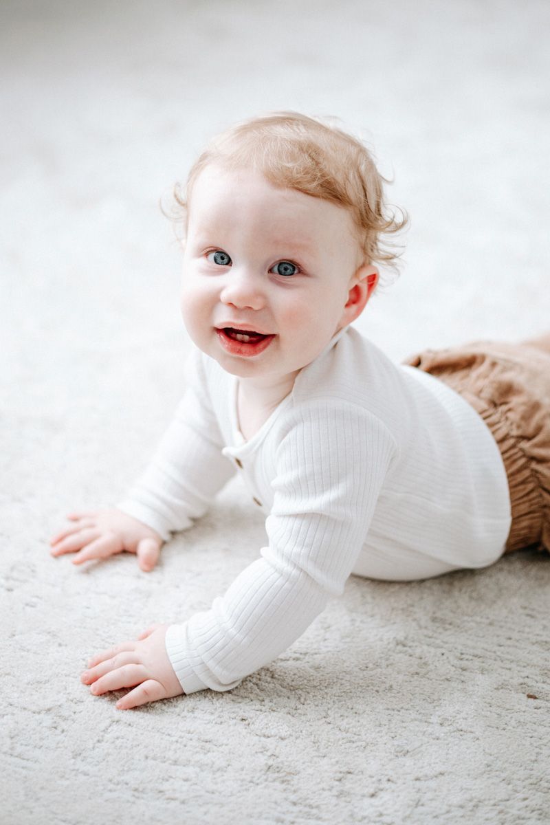 Smiling baby in white ribbed outfit crawls on light carpet during portrait session.