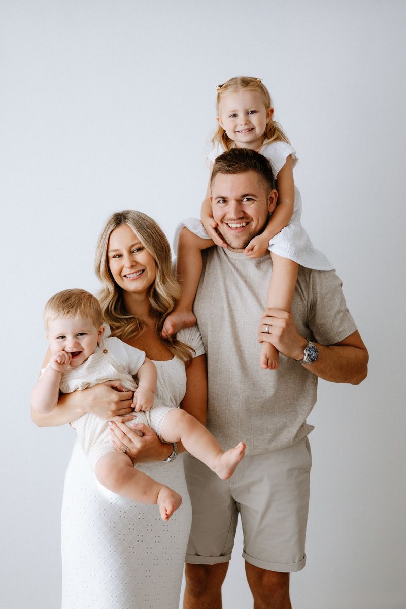 Happy family of four posing together, parents holding two young children, wearing neutral tones against white background.