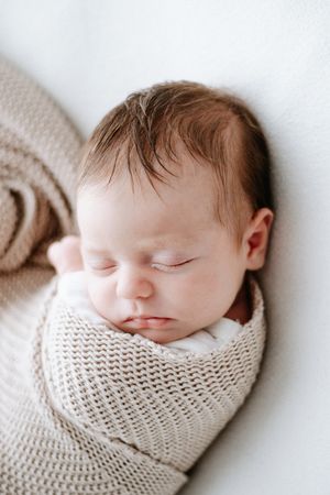 A sleeping newborn baby wrapped snugly in a cream-colored knit blanket against a white background.
