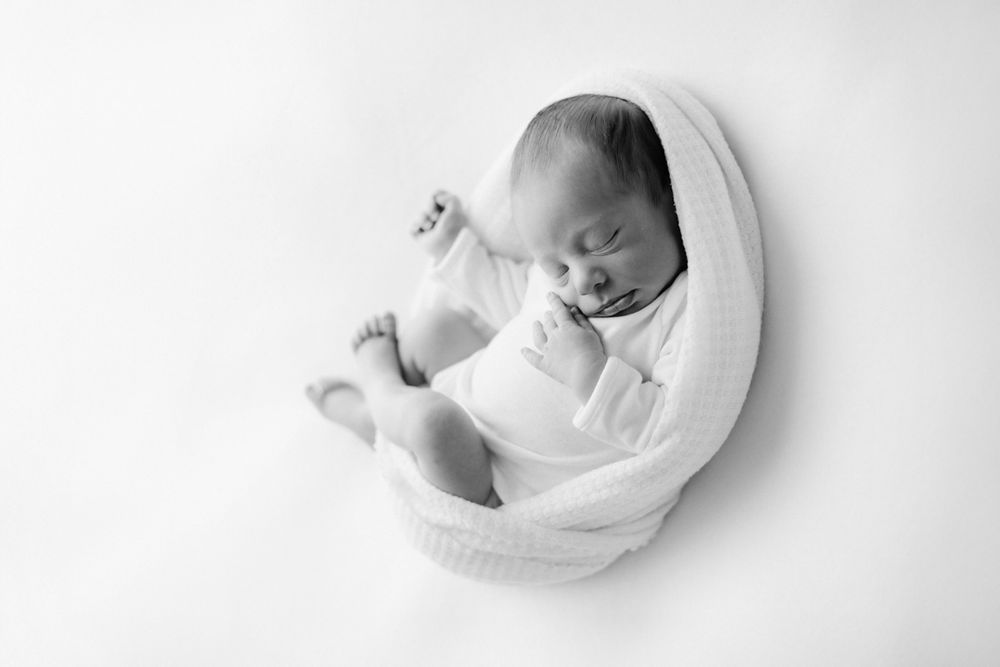 Black and white newborn photo of sleeping baby wrapped in white knit blanket, curled in fetal position on white background.