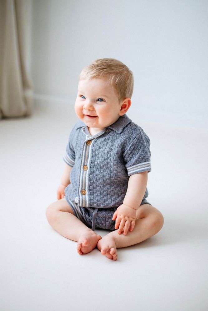 Baby wearing grey patterned shirt and shorts sits on white floor while smiling upward at milestone photoshoot in nottingham