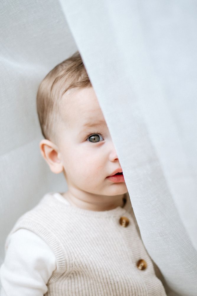 A young toddler in a cream sweater peeks out from behind a white curtain at milestone photoshoot in mansfield