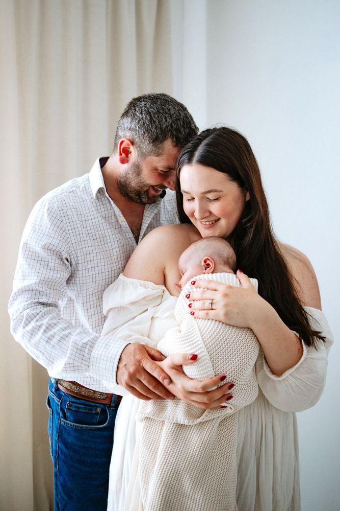 Parents share tender moment while holding their newborn baby wrapped in white against light curtained background.