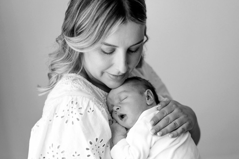 A tender black and white portrait of a parent in a white embroidered top holding a sleeping newborn baby.