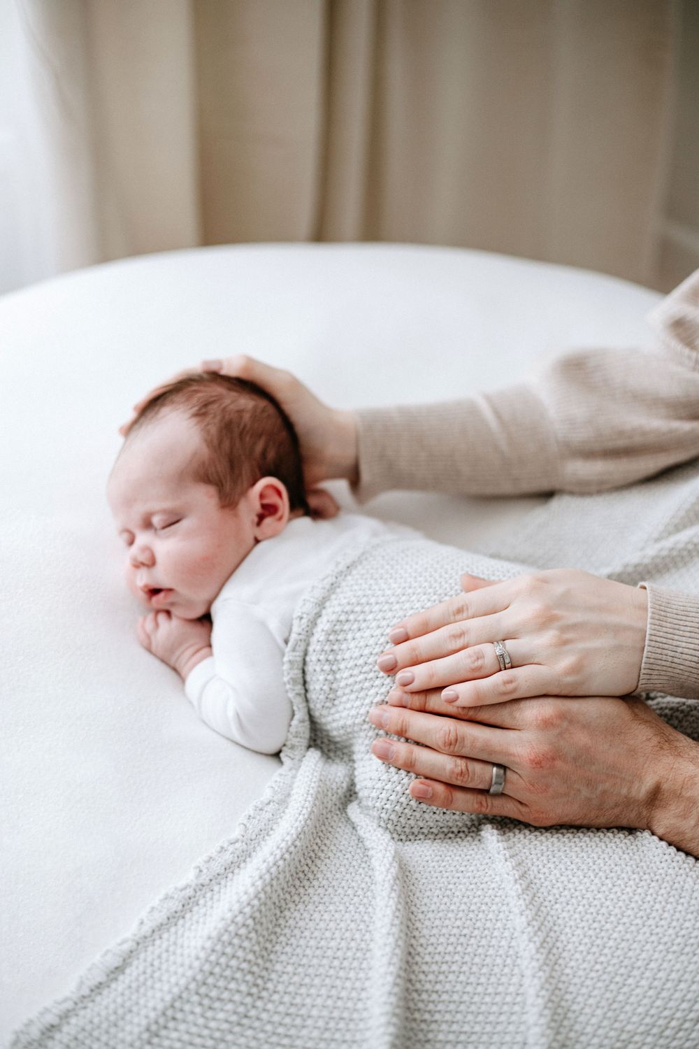 A sleeping baby wrapped in a knitted white blanket while hands lovingly rest on their back.
