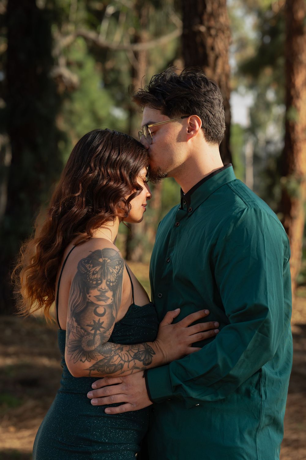 A romantic couple in green and gray share tender moments together in a wooded outdoor setting at sunset.