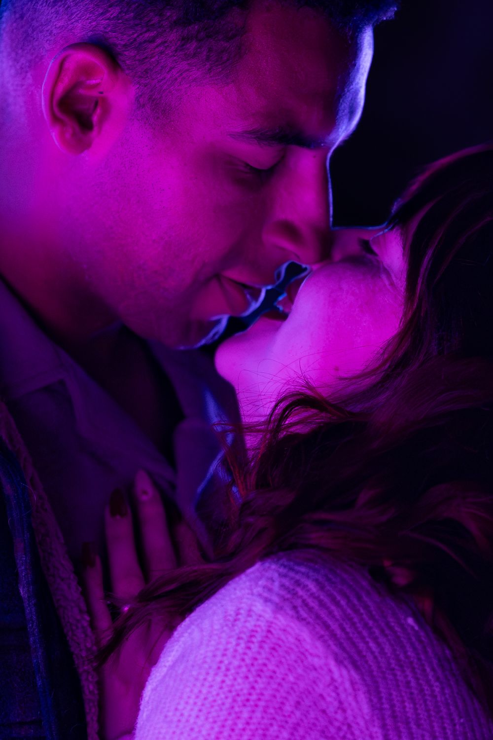 A romantic couple in purple lighting share an intimate moment wearing white knit clothing.