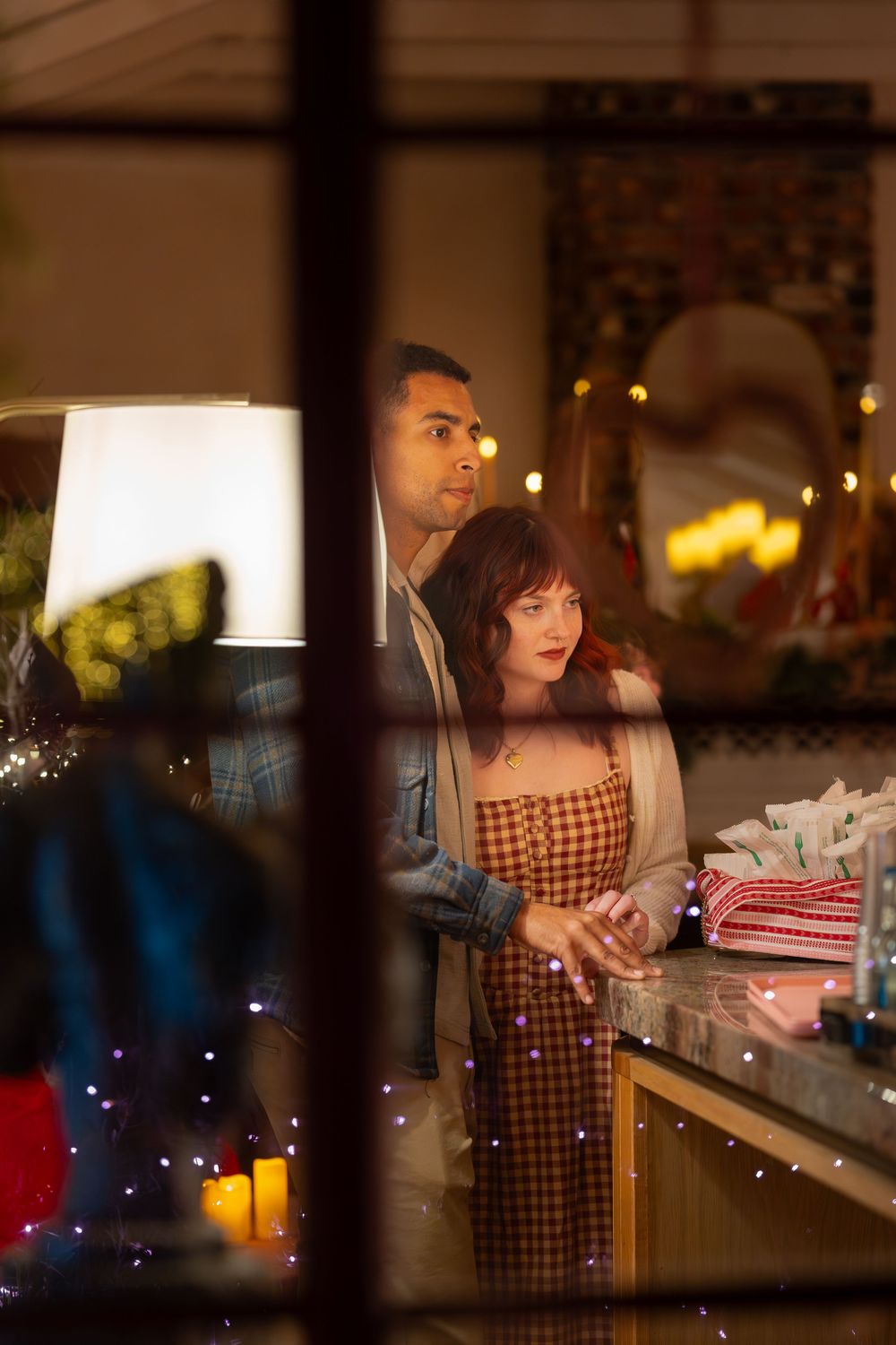 A couple stands at a dimly lit counter with candles and festive lights creating a romantic evening atmosphere.