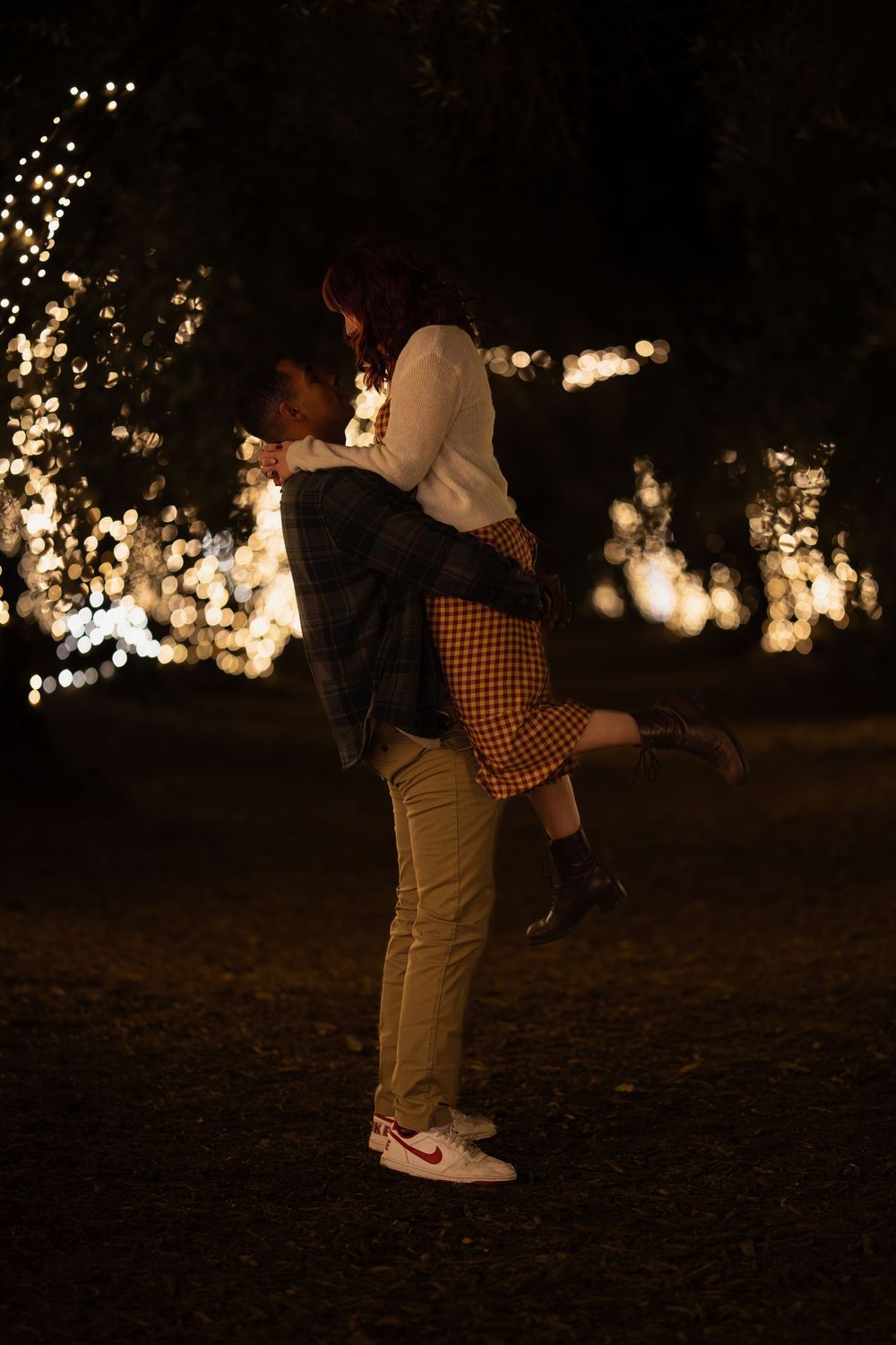 A romantic couple embraces in silhouette against twinkling lights and trees at dusk during an outdoor photoshoot.