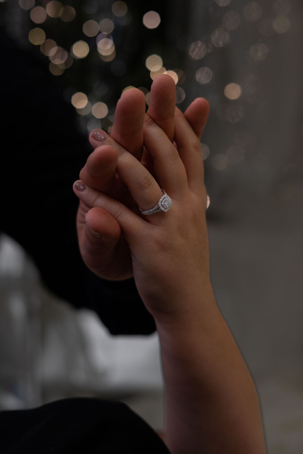 A close-up view of hands with an engagement ring against a blurred bokeh background.