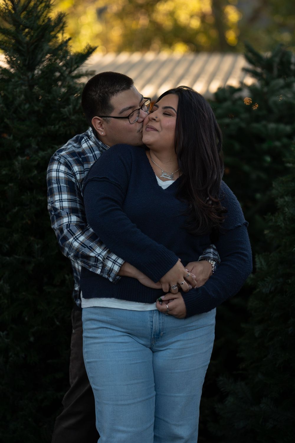 A couple in matching light jeans and dark tops share tender moments against an evergreen backdrop at sunset.