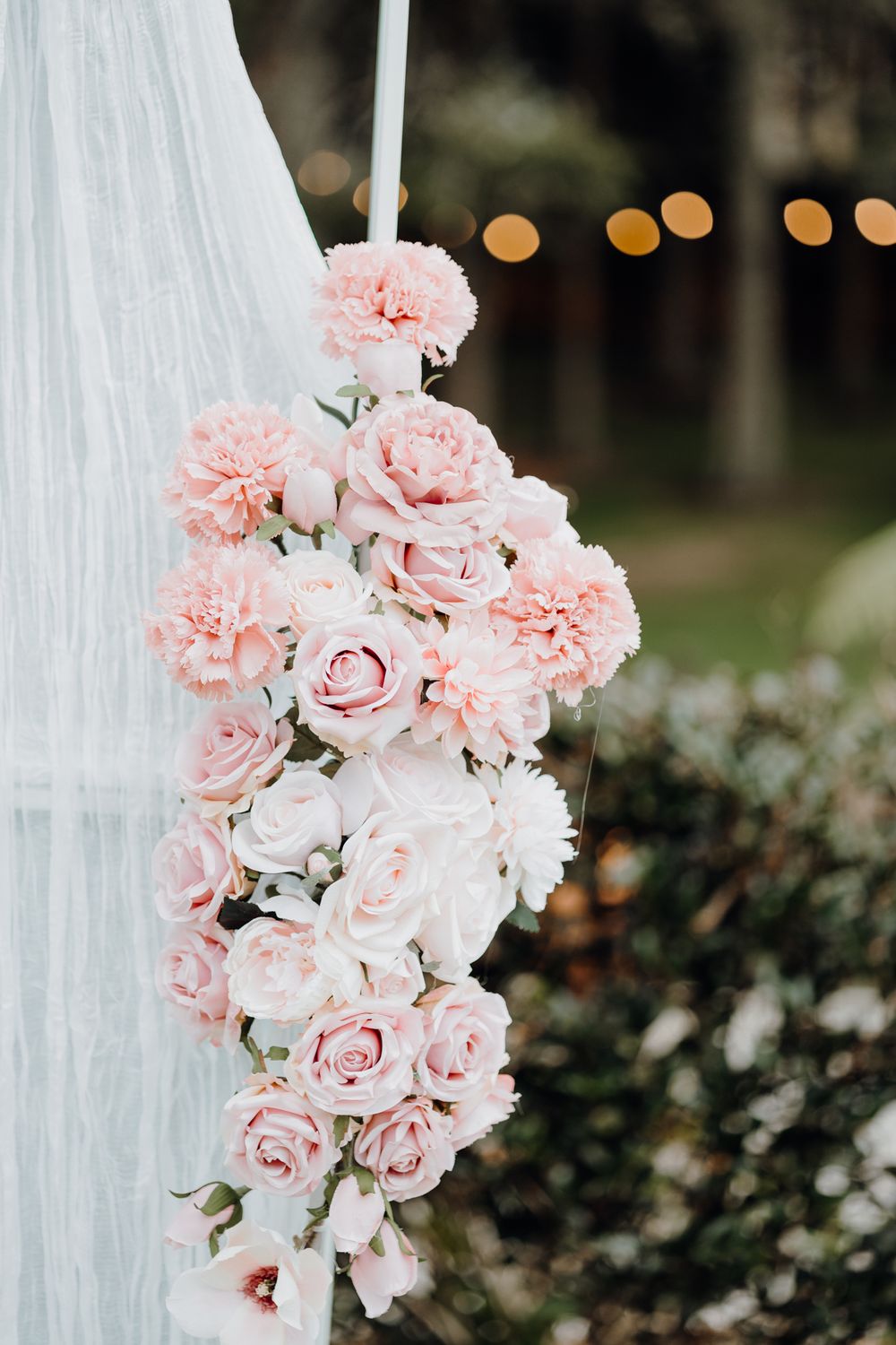 Close-up of delicate pink roses and carnations arranged on white fabric with bokeh lights in background.