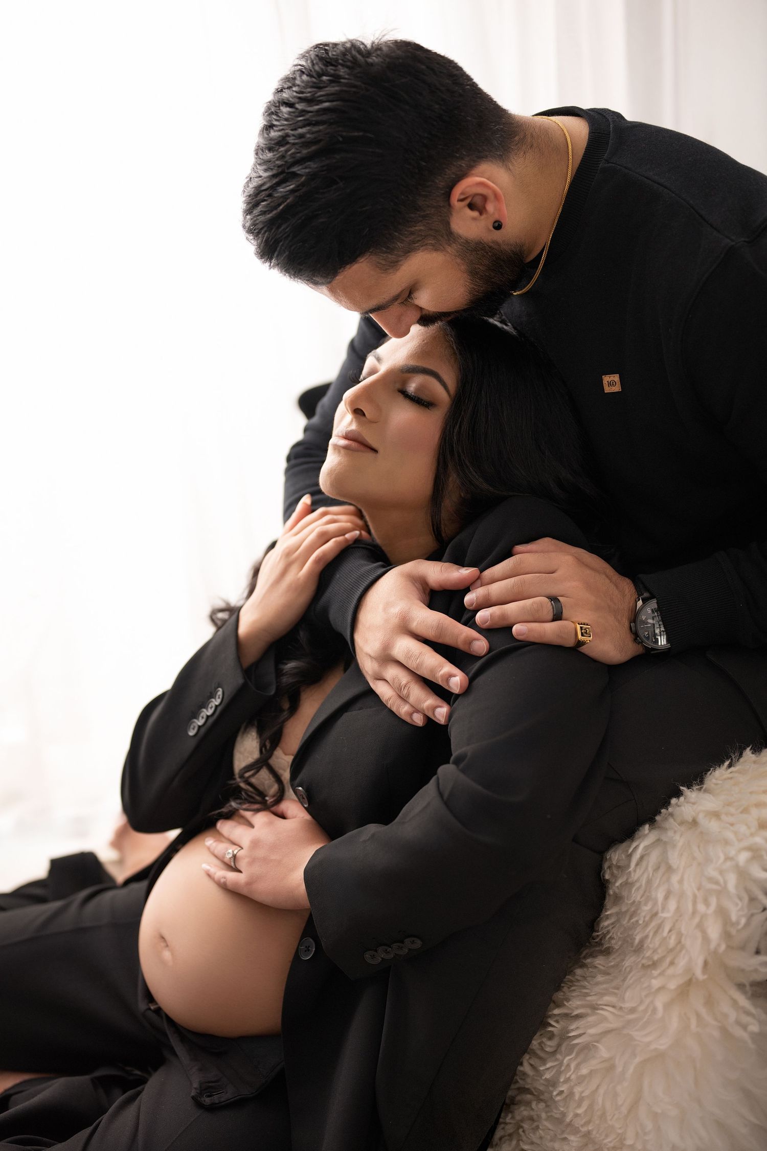 Intimate maternity photo session showing couple embracing in black formal wear against white backdrop.