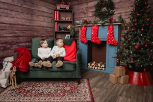 Two children sit on a green couch in a cozy Christmas setting with stockings and decorations.