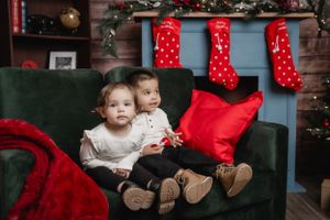 Two small children sit together on a dark green couch with red Christmas stockings hanging behind them.
