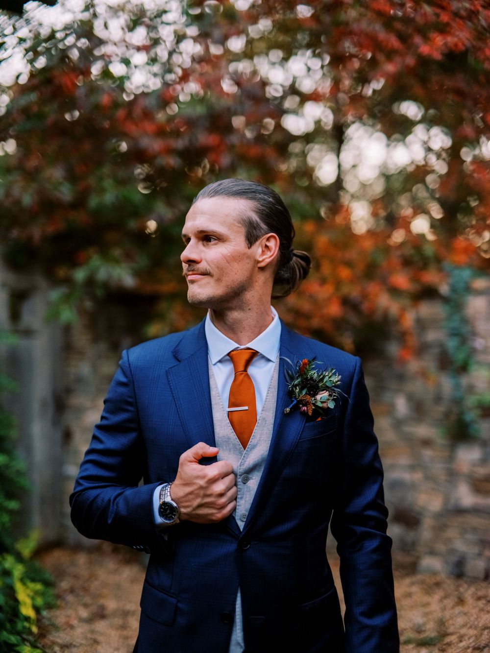 Groom in navy suit with orange tie poses against autumn foliage backdrop.