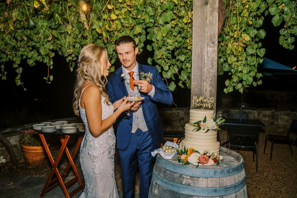 Newlyweds share cake cutting moment at rustic outdoor reception with wine barrel decor.