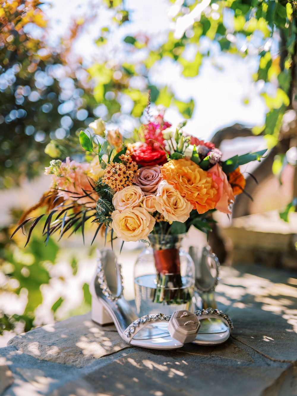 A beautiful fall wedding bouquet with orange and pink roses sits next to bridal shoes in natural outdoor lighting.