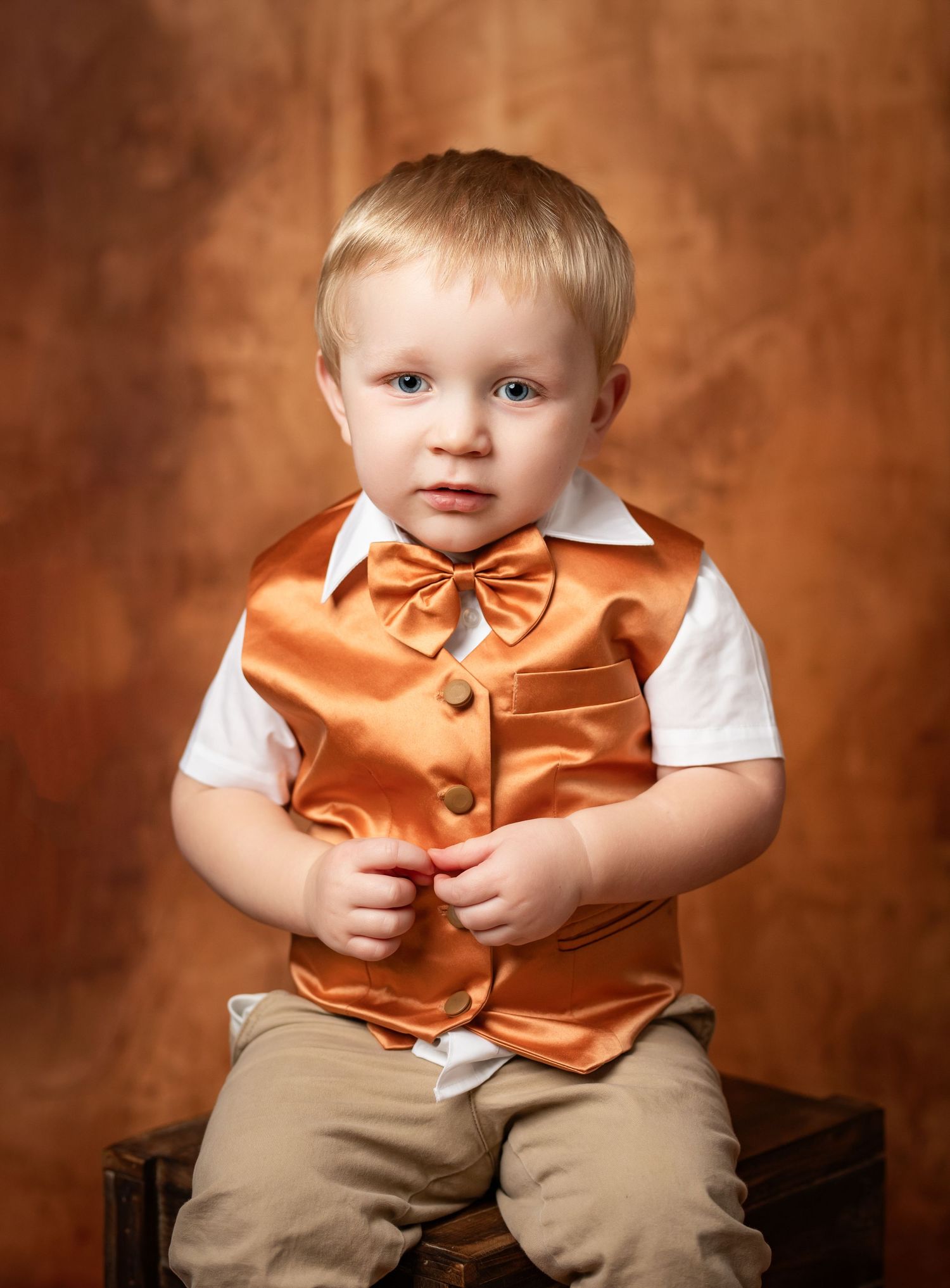 Portrait of a young child in an orange satin vest and bowtie with white shirt sitting against a brown backdrop.